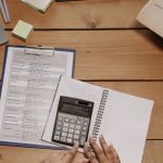 Top view of hands using calculator on office desk surrounded by documents and sticky notes.